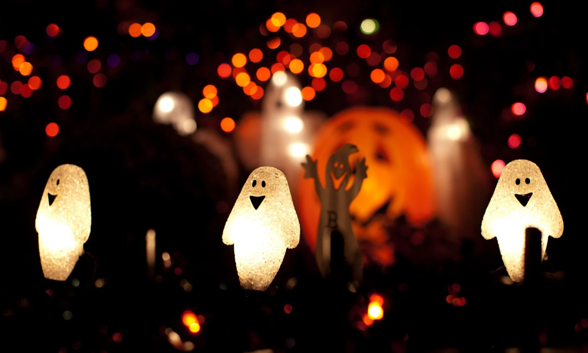 Small, friendly glowing ghost figures light up a dark background decorated with warm orange and red bokeh lights.