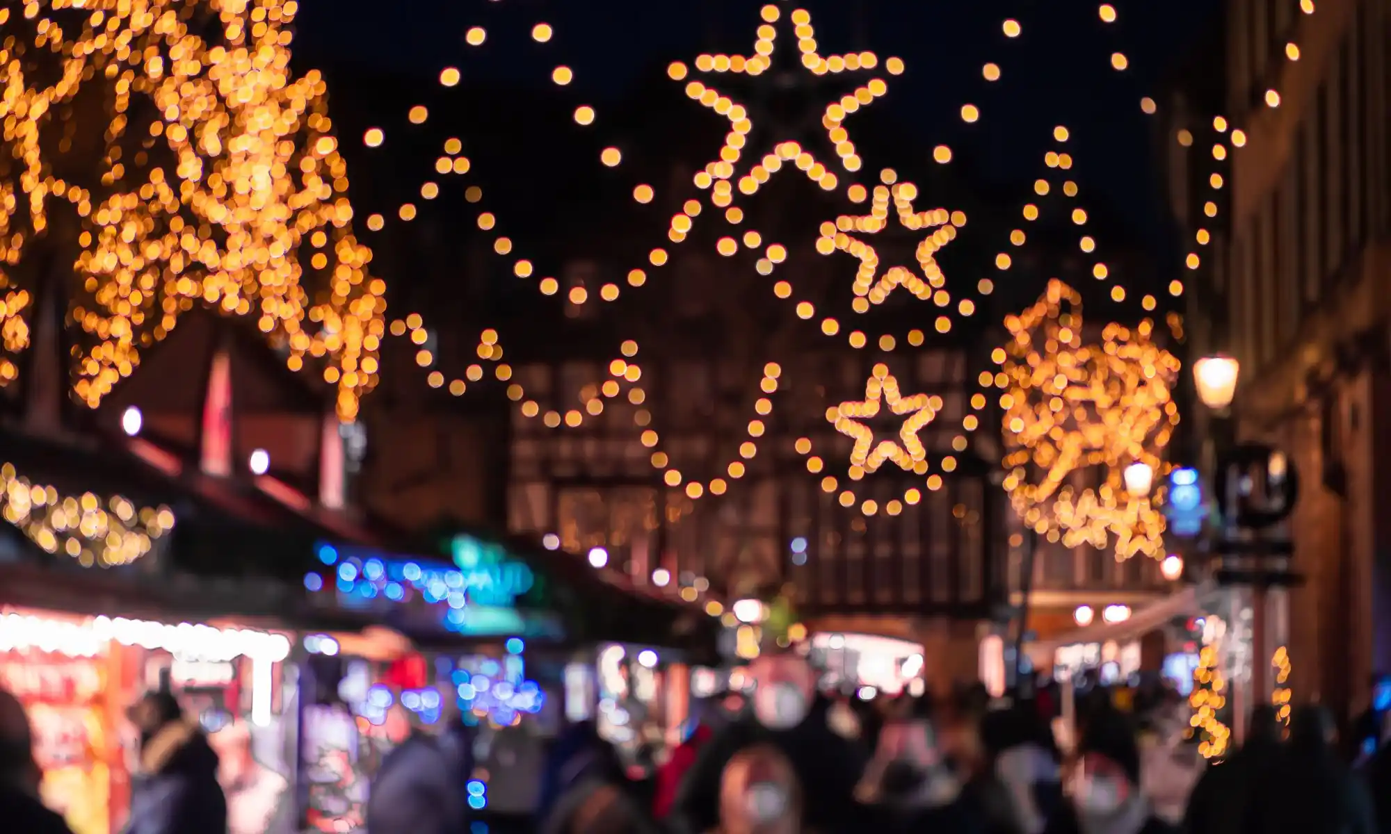 Star-shaped holiday lights glow above blurred outdoor market stalls and pedestrians at night.